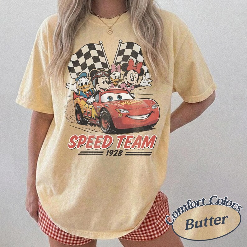 K&ouml;nnte beinhalten: Hellgelbes T-Shirt mit einer Grafik von Disney-Figuren in einem roten Rennwagen. Das Design umfasst Micky Maus, Donald Duck, Daisy Duck und Minnie Maus. Der Text "SPEED TEAM 1928" befindet sich unter dem Auto. Zwei karierte Flaggen sind im Hintergrund.