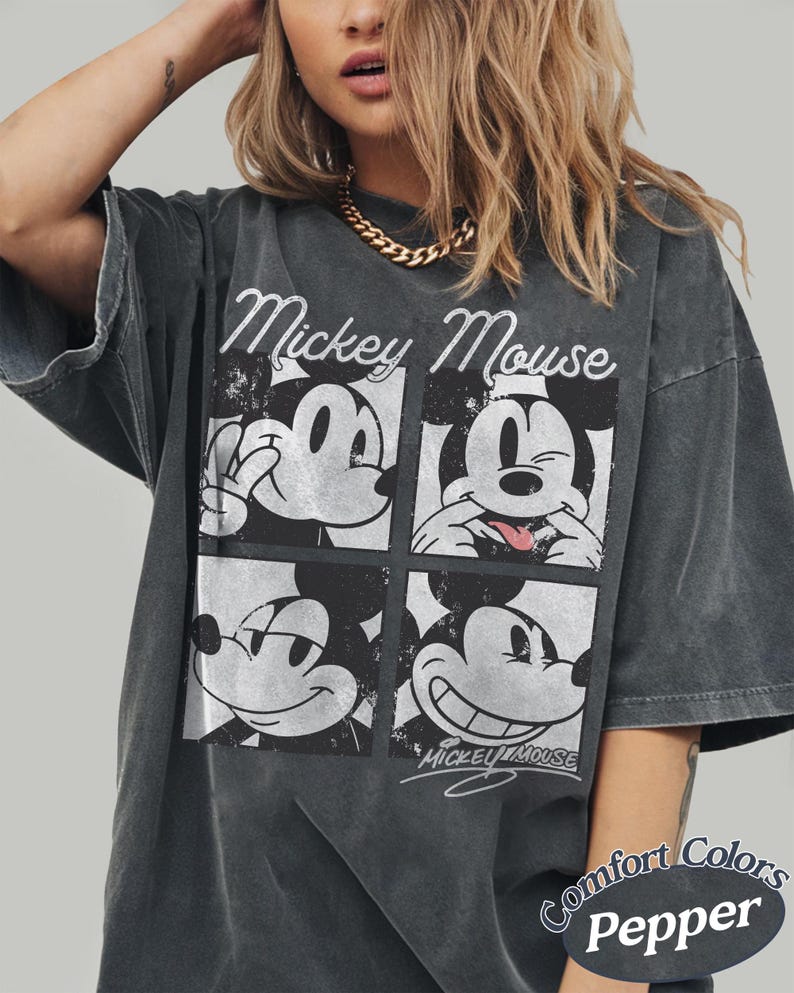 Op de afbeelding: Donkergrijs T-shirt met een vierpaneelsafbeelding van Mickey Mouse met verschillende uitdrukkingen. De woorden "Mickey Mouse" staan in het wit boven de afbeelding. Het shirt is gelabeld met "Comfort Colors Pepper".