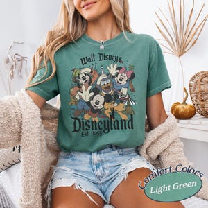 May include: Light green t-shirt featuring a vintage-style graphic of Disney characters, including Mickey Mouse, Donald Duck, and Minnie Mouse. The text "Walt Disney's Disneyland Est. 1955" is printed above and below the characters. The shirt is labeled "Comfort Colors Light Green".