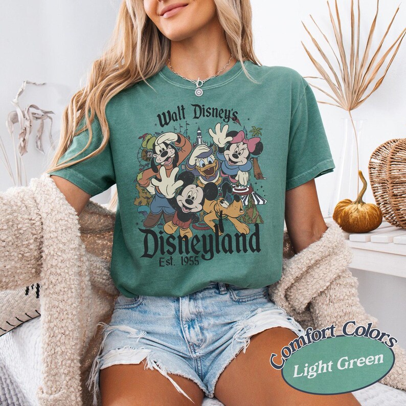 May include: Light green t-shirt featuring a vintage-style graphic of Disney characters, including Mickey Mouse, Donald Duck, and Minnie Mouse. The text "Walt Disney's Disneyland Est. 1955" is printed above and below the characters. The shirt is labeled "Comfort Colors Light Green".