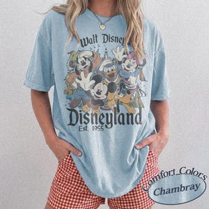 May include: Light blue t-shirt featuring a vintage Disneyland graphic with Mickey Mouse, Donald Duck, Goofy, and Minnie Mouse. The shirt has the text "Walt Disney Disneyland Est. 1955". The shirt is a Comfort Colors Chambray.