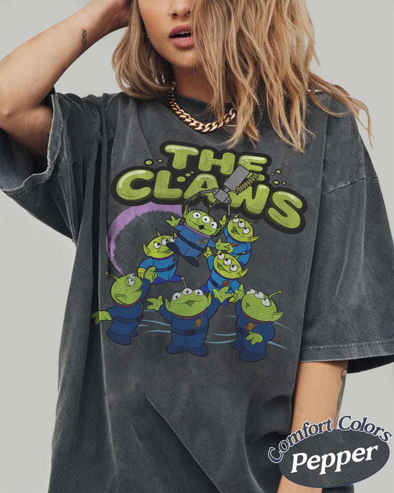 May include: Dark gray t-shirt featuring a graphic of green aliens in blue uniforms with the text "THE CLAWS". The design includes a purple arc and a claw machine. The shirt is from Comfort Colors.