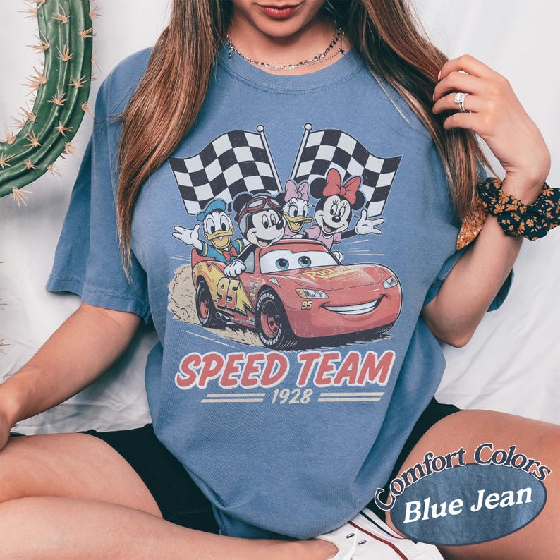 K&ouml;nnte beinhalten: Blaues Comfort Colors T-Shirt mit einem Aufdruck von Disney-Figuren: Mickey Mouse, Donald Duck, Daisy Duck und Minnie Mouse in einem roten Rennwagen mit karierten Flaggen. Der Text "SPEED TEAM 1928" ist darunter gedruckt.