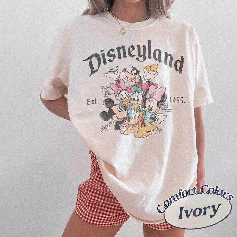 May include: Ivory-colored t-shirt with a Disneyland graphic featuring Mickey Mouse, Minnie Mouse, Donald Duck, Daisy Duck, Goofy, and Pluto. The shirt includes the text "Disneyland Est. 1955." and is a Comfort Colors brand.