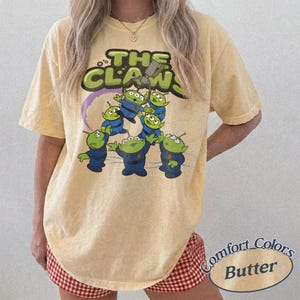 May include: Butter-colored t-shirt featuring a graphic of the aliens from the movie Toy Story. The design includes the text "THE CLAW" in green and yellow, with the aliens in blue uniforms. The shirt is a Comfort Colors brand.