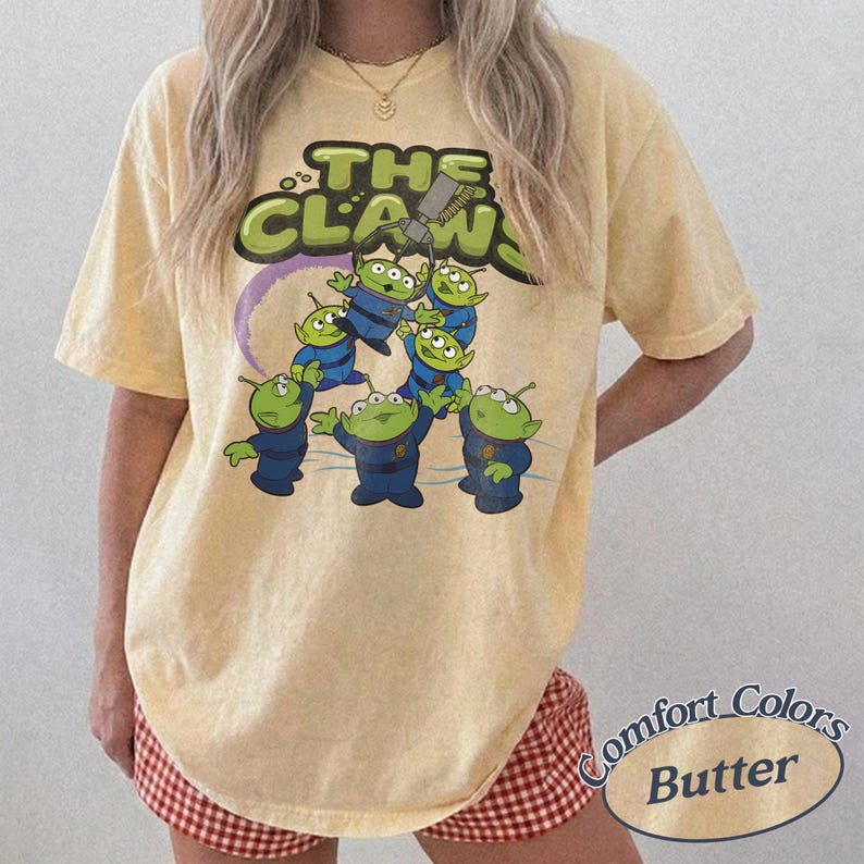 May include: Butter-colored t-shirt featuring a graphic of the aliens from the movie Toy Story. The design includes the text "THE CLAW" in green and yellow, with the aliens in blue uniforms. The shirt is a Comfort Colors brand.