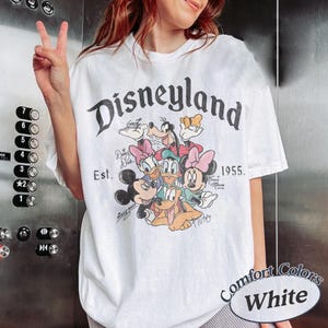 May include: White t-shirt with a Disneyland graphic featuring Mickey Mouse, Minnie Mouse, Donald Duck, Daisy Duck, Goofy, and Pluto. The shirt includes the text "Disneyland" and "Est. 1955." The shirt is labeled "Comfort Colors White."