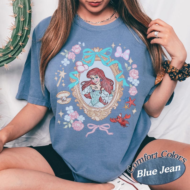 Puede incluir: Camiseta azul vaquero Comfort Colors con una ilustraci&oacute;n enmarcada de Ariel de La Sirenita. El dise&ntilde;o incluye rosas rosas, lazos azules, una concha, un cangrejo rojo y una estrella de mar. La camiseta tiene mangas cortas.
