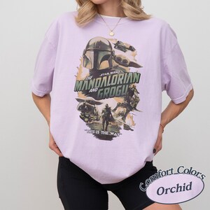 Może przedstawiać: Jasnofioletowa koszulka z grafiką Star Wars z napisem "The Mandalorian and Grogu". Projekt zawiera postacie z serialu i frazę "This is the Way". Koszulka marki Comfort Colors, kolor "Orchid".