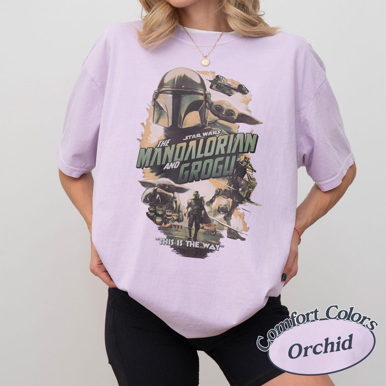 Może przedstawiać: Jasnofioletowa koszulka z grafiką Star Wars z napisem "The Mandalorian and Grogu". Projekt zawiera postacie z serialu i frazę "This is the Way". Koszulka marki Comfort Colors, kolor "Orchid".