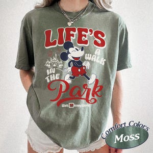 May include: Moss green t-shirt featuring Mickey Mouse and the text "Life's a Walk in the Park" in red and white. The design includes a castle graphic and "Walt Disney World" text. The shirt is a Comfort Colors brand.