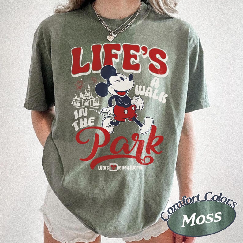 May include: Moss green t-shirt featuring Mickey Mouse and the text "Life's a Walk in the Park" in red and white. The design includes a castle graphic and "Walt Disney World" text. The shirt is a Comfort Colors brand.