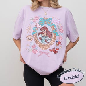 Puede incluir: Una camiseta lila con una ilustraci&oacute;n enmarcada de una mujer pelirroja con un vestido azul, rodeada de elementos florales y n&aacute;uticos. La camiseta tiene el texto "Comfort Colors Orchid" en un dise&ntilde;o circular.