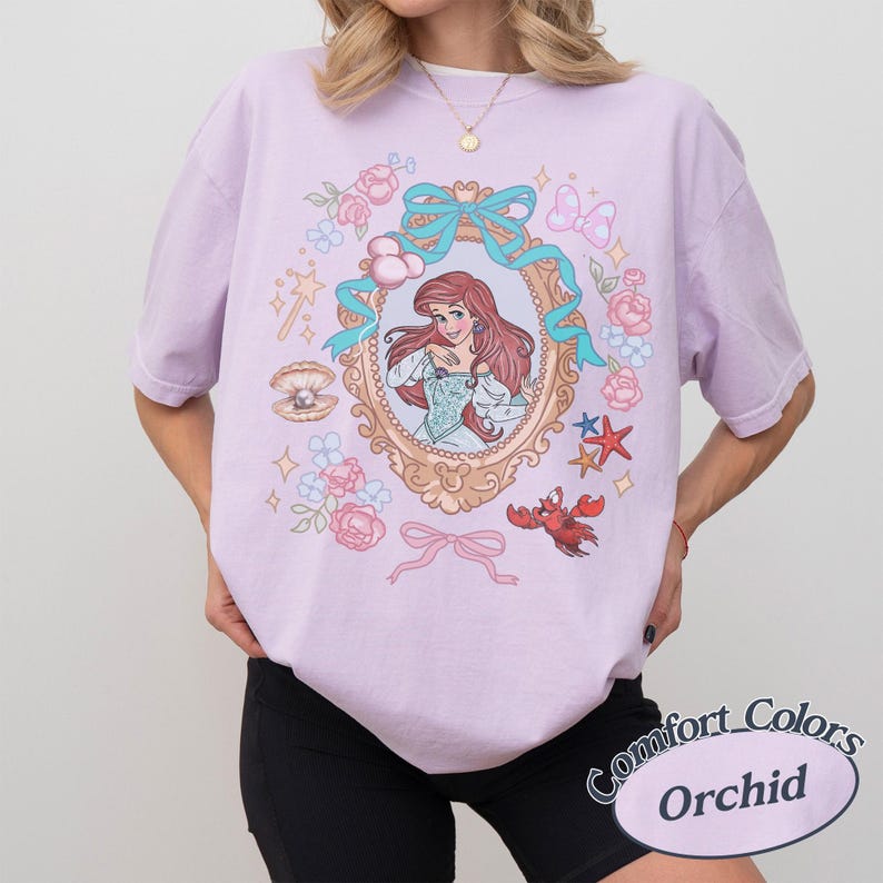 Puede incluir: Una camiseta lila con una ilustraci&oacute;n enmarcada de una mujer pelirroja con un vestido azul, rodeada de elementos florales y n&aacute;uticos. La camiseta tiene el texto "Comfort Colors Orchid" en un dise&ntilde;o circular.