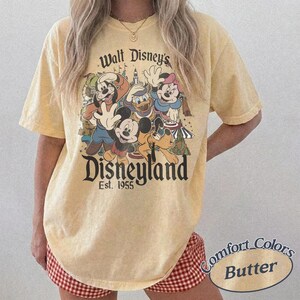 May include: A light yellow t-shirt featuring a graphic of Disney characters, including Mickey Mouse, Donald Duck, and Goofy, with the text "Walt Disney's Disneyland Est. 1955". The shirt is labeled "Comfort Colors Butter".