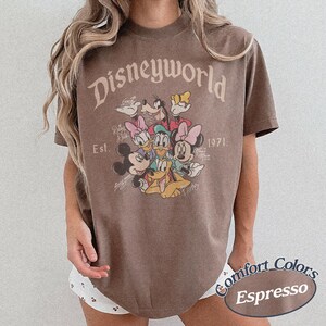 Może przedstawiać: Brązowa koszulka w kolorze espresso z grafiką Disneyworld z Myszką Miki, Myszka Minnie, Kaczorem Donaldem, Daisy, Goofym i Plutonem. Koszulka zawiera napis "Disneyworld" i "Est. 1971". Koszulka marki Comfort Colors.