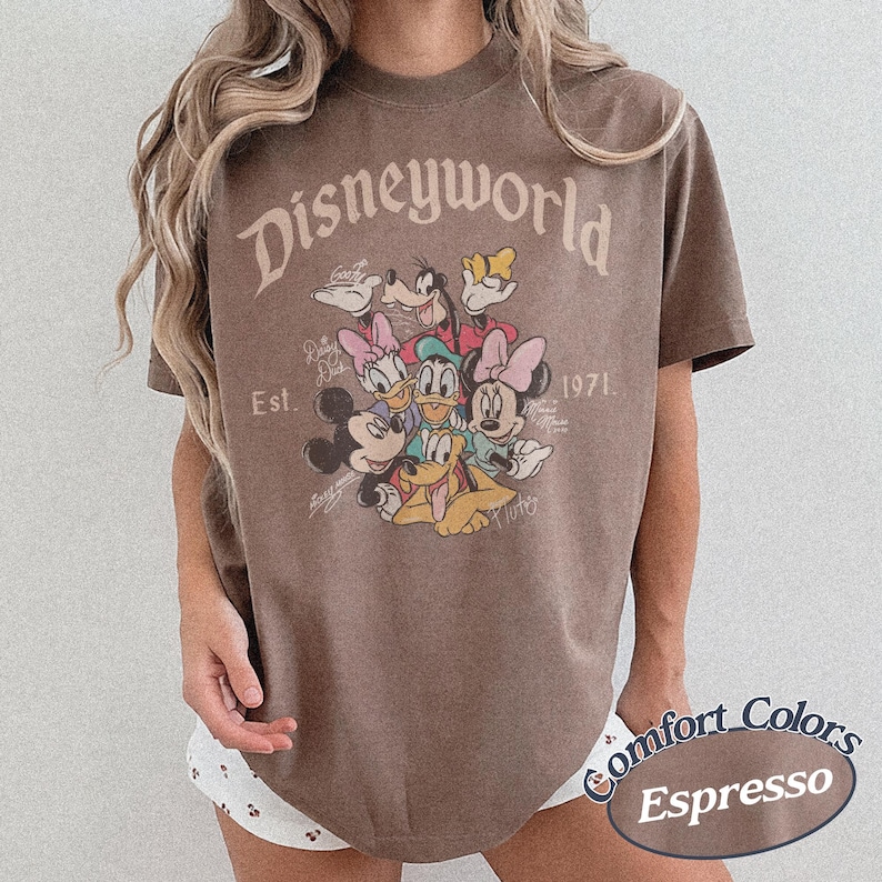 Może przedstawiać: Brązowa koszulka w kolorze espresso z grafiką Disneyworld z Myszką Miki, Myszka Minnie, Kaczorem Donaldem, Daisy, Goofym i Plutonem. Koszulka zawiera napis "Disneyworld" i "Est. 1971". Koszulka marki Comfort Colors.
