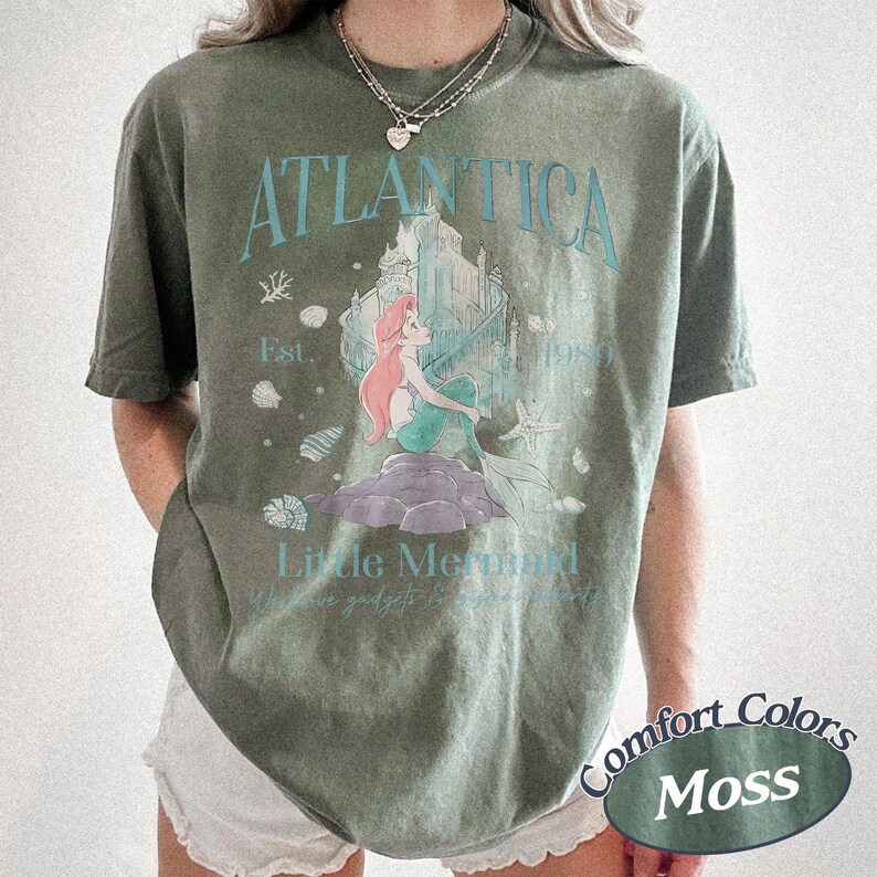 Może przedstawiać: Zielona koszulka w kolorze mchu z grafiką Arielki z Małej Syrenki, z napisem "Atlantica" i "Little Mermaid". Projekt zawiera zamek i muszle. Koszulka jest oznaczona jako "Comfort Colors Moss".