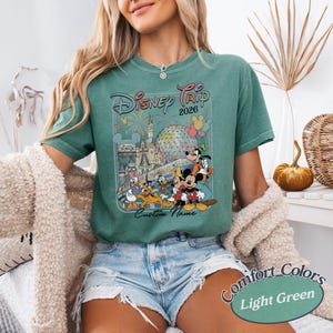 May include: Light green t-shirt with a Disney Trip 2026 graphic featuring Mickey Mouse, Donald Duck, and other characters. The shirt has the words "Custom Name" below the graphic and "Comfort Colors Light Green" at the bottom.
