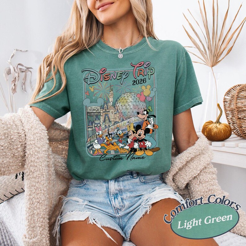 May include: Light green t-shirt with a Disney Trip 2026 graphic featuring Mickey Mouse, Donald Duck, and other characters. The shirt has the words "Custom Name" below the graphic and "Comfort Colors Light Green" at the bottom.