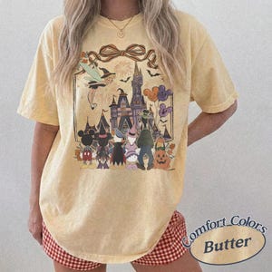 Pode incluir: Camiseta amarela manteiga com um gr&aacute;fico tem&aacute;tico de Halloween. O design inclui um castelo, personagens, morcegos e fogos de artif&iacute;cio. A camisa tem um ajuste folgado e mangas curtas. O texto "Comfort Colors Butter" &eacute; impresso no canto inferior direito.