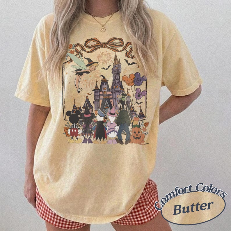 Pode incluir: Camiseta amarela manteiga com um gr&aacute;fico tem&aacute;tico de Halloween. O design inclui um castelo, personagens, morcegos e fogos de artif&iacute;cio. A camisa tem um ajuste folgado e mangas curtas. O texto "Comfort Colors Butter" &eacute; impresso no canto inferior direito.