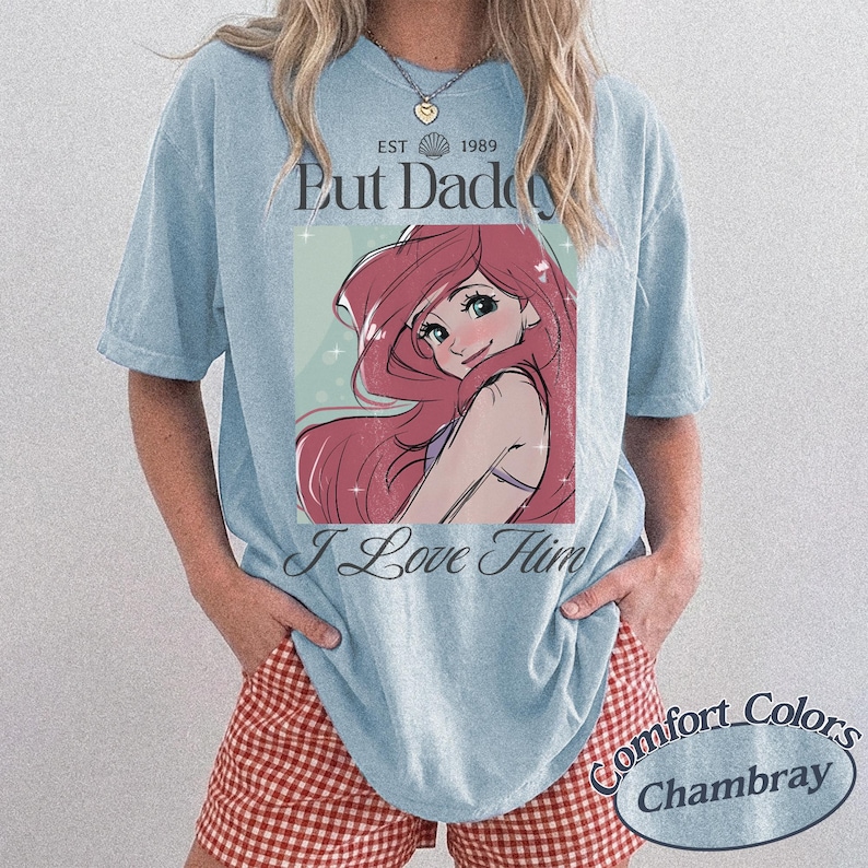 K&ouml;nnte beinhalten: Hellblaues T-Shirt mit einer Cartoon-Illustration einer Figur mit roten Haaren und dem Text "But Daddy I Love Him". Das Shirt hat auch den Text "EST 1989" und "Comfort Colors Chambray".