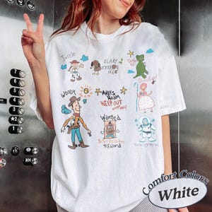 Puede incluir: Camiseta blanca con ilustraciones de dibujos animados coloridos de personajes y texto de la pel&iacute;cula Toy Story. La camiseta incluye las palabras "Woody", "Jessie", "Slinky", "Andy's Room Keep Out" y "Buzz Lightyear". La camiseta tambi&eacute;n tiene las palabras "Comfort Colors White".