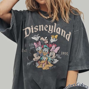 May include: Dark gray t-shirt featuring the word "Disneyland" above a graphic of Disney characters. The shirt includes the text "Est. 1955" and the Comfort Colors Pepper label.