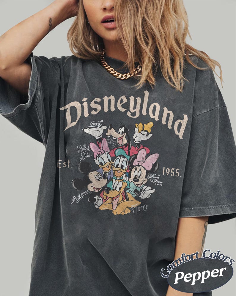 May include: Dark gray t-shirt featuring the word "Disneyland" above a graphic of Disney characters. The shirt includes the text "Est. 1955" and the Comfort Colors Pepper label.