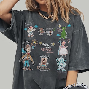 Puede incluir: Camiseta gris oscuro con ilustraciones de dibujos animados de personajes de Toy Story, incluyendo a Woody, Jessie y Slinky. El dise&ntilde;o presenta el texto "ANDY'S ROOM KEEP OUT" y "$50 Billion Reward". La camiseta est&aacute; etiquetada como "Comfort Colors Pepper".