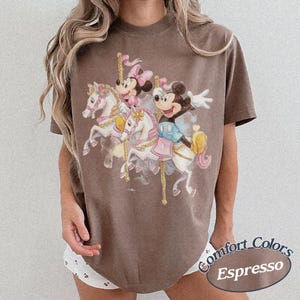 Puede incluir: Camiseta marr&oacute;n con un dise&ntilde;o de carrusel que presenta a Mickey y Minnie Mouse montados en caballos blancos. El dise&ntilde;o incluye detalles en rosa, azul y amarillo. La camiseta est&aacute; etiquetada como "Comfort Colors" y "Espresso".