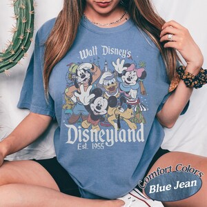 May include: A blue jean colored t-shirt featuring a graphic of Disney characters including Mickey Mouse, Minnie Mouse, Donald Duck, and Goofy. The text "Walt Disney's Disneyland Est. 1955" is printed on the front. The shirt is a comfort color.