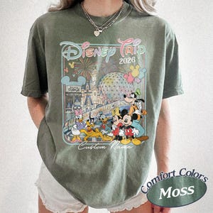 May include: An olive green t-shirt with a Disney Trip 2026 design, showcasing Mickey Mouse, Donald Duck, Goofy, and other characters. The graphic includes fireworks, a castle, and "Custom Name". The shirt is labelled "Comfort Colors Moss".
