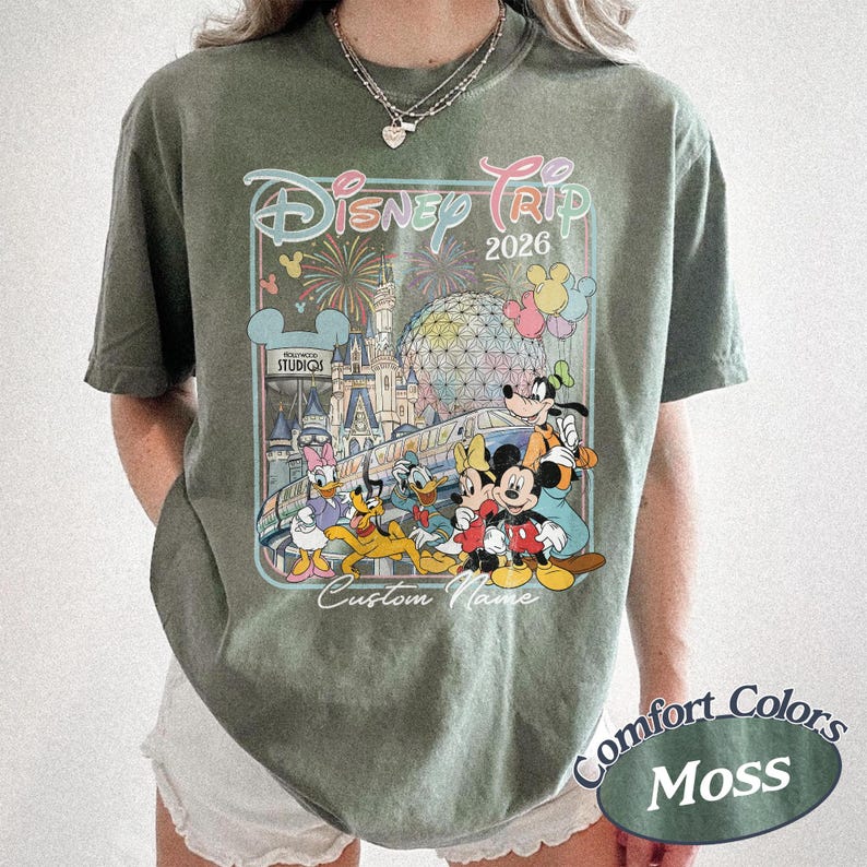 May include: An olive green t-shirt with a Disney Trip 2026 design, showcasing Mickey Mouse, Donald Duck, Goofy, and other characters. The graphic includes fireworks, a castle, and "Custom Name". The shirt is labelled "Comfort Colors Moss".