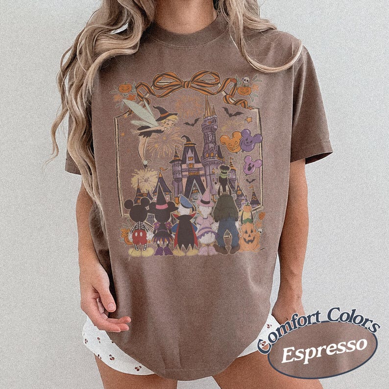 Pode incluir: Camiseta cor espresso com um gr&aacute;fico tem&aacute;tico de Halloween. O design inclui personagens de desenhos animados, um castelo e uma fada. A camiseta tem gola redonda e mangas curtas. As palavras "Comfort Colors" e "Espresso" s&atilde;o impressas na parte inferior direita.