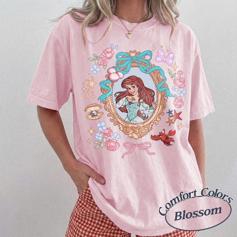 Puede incluir: Camiseta rosa claro con una ilustraci&oacute;n enmarcada de un personaje de dibujos animados con pelo rojo y un vestido azul. El marco est&aacute; rodeado de flores y lazos rosas y azules. El texto "Comfort Colors Blossom" est&aacute; impreso en la parte inferior derecha.