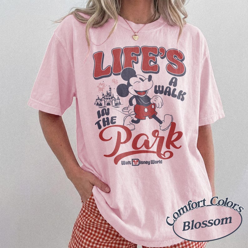 May include: A light pink t-shirt with a graphic of Mickey Mouse walking in front of a castle, with the text "LIFE'S A WALK IN THE PARK" in red and white. The shirt also has the text "Walt Disney World" and "Comfort Colors Blossom".