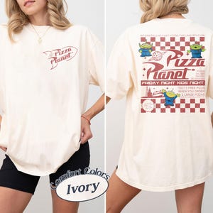 May include: Ivory-colored t-shirt with the Pizza Planet logo on the front and back. The back features the Pizza Planet logo, aliens, and text. The shirt is a Comfort Colors brand.