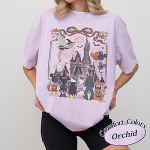 Pode incluir: Uma camiseta lil&aacute;s clara com um gr&aacute;fico da Disney com tema de Halloween. O design inclui Mickey Mouse, Pato Donald e uma ab&oacute;bora, com um castelo ao fundo. A camisa tem as palavras "Comfort Colors" e "Orchid" impressas.