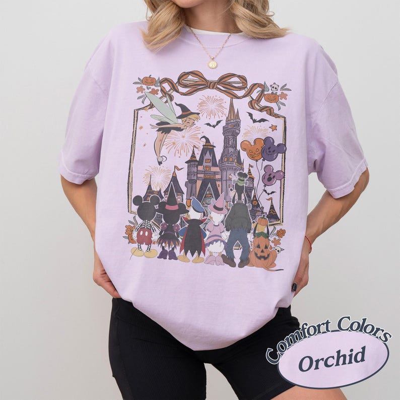 Pode incluir: Uma camiseta lil&aacute;s clara com um gr&aacute;fico da Disney com tema de Halloween. O design inclui Mickey Mouse, Pato Donald e uma ab&oacute;bora, com um castelo ao fundo. A camisa tem as palavras "Comfort Colors" e "Orchid" impressas.