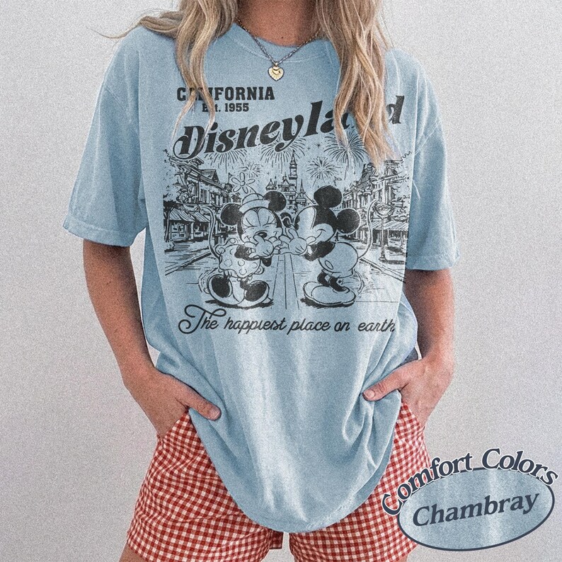 Puede incluir: Camiseta azul claro con un gr&aacute;fico en blanco y negro de Mickey y Minnie Mouse frente a Disneyland. La camiseta tiene el texto "California Est. 1955 Disneyland The happiest place on earth". La camiseta es Comfort Colors Chambray.