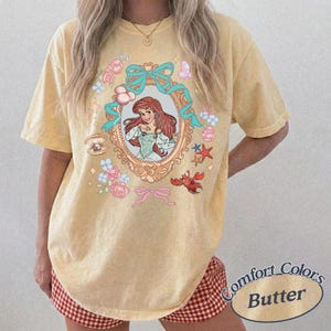 Puede incluir: Camiseta de color mantequilla con una ilustraci&oacute;n enmarcada de una mujer pelirroja con un vestido, rodeada de elementos florales y acu&aacute;ticos. El dise&ntilde;o incluye un cangrejo, estrellas de mar y lazos. La camiseta tiene las palabras "Comfort Colors Butter".