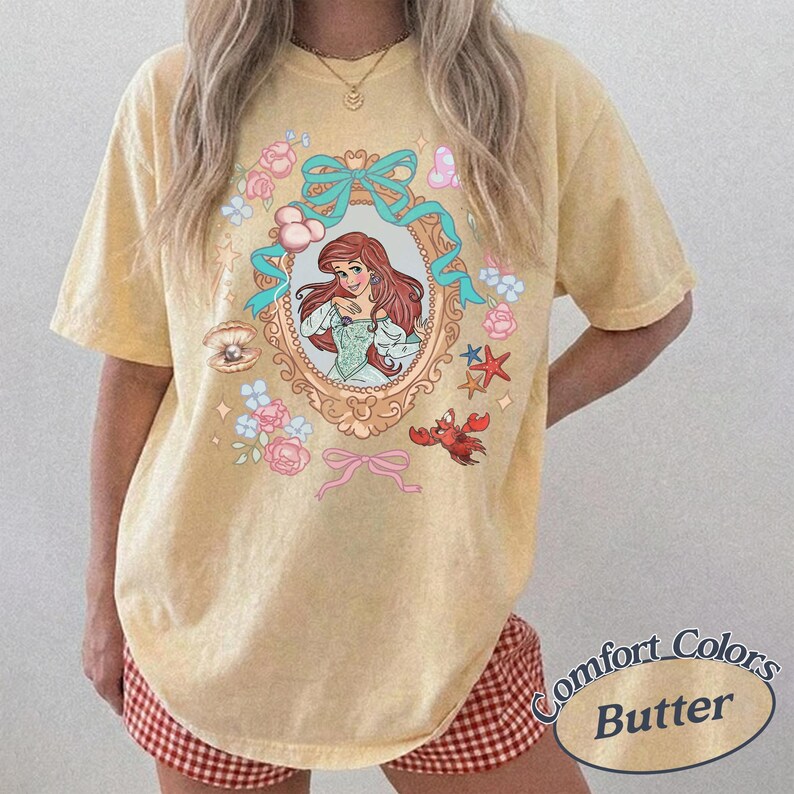 Puede incluir: Camiseta de color mantequilla con una ilustraci&oacute;n enmarcada de una mujer pelirroja con un vestido, rodeada de elementos florales y acu&aacute;ticos. El dise&ntilde;o incluye un cangrejo, estrellas de mar y lazos. La camiseta tiene las palabras "Comfort Colors Butter".