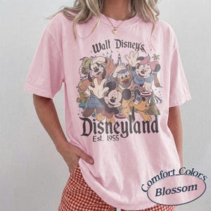 May include: A light pink t-shirt featuring a graphic of Disney characters including Mickey Mouse, Donald Duck, and Goofy. The shirt has the text "Walt Disney's Disneyland Est. 1955" and "Comfort Colors Blossom".