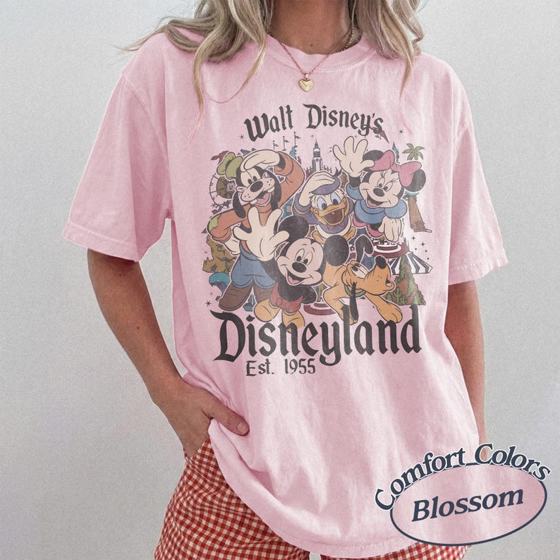 May include: A light pink t-shirt featuring a graphic of Disney characters including Mickey Mouse, Donald Duck, and Goofy. The shirt has the text "Walt Disney's Disneyland Est. 1955" and "Comfort Colors Blossom".