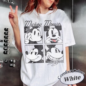 Op de afbeelding: Wit T-shirt met een zwart-wit Mickey Mouse-afbeelding in vier verschillende poses. Het shirt heeft de woorden "Mickey Mouse" boven de afbeeldingen en "Mickey Mouse" in script eronder. Het shirt heeft ook de woorden "Comfort Colors White" rechtsonder.