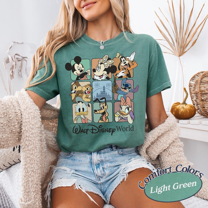 May include: Light green t-shirt featuring a grid of Disney characters including Mickey Mouse, Donald Duck, and Stitch. The shirt has the text "Walt Disney World" and "Comfort Colors" printed on it.