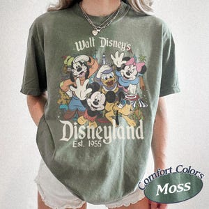 May include: A moss green t-shirt featuring a graphic of Disney characters including Mickey Mouse, Donald Duck, and Goofy. The text "Walt Disney's Disneyland Est. 1955" is printed above the characters. The shirt also has the text "Comfort Colors Moss".