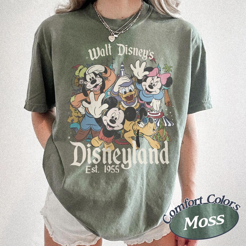 May include: A moss green t-shirt featuring a graphic of Disney characters including Mickey Mouse, Donald Duck, and Goofy. The text "Walt Disney's Disneyland Est. 1955" is printed above the characters. The shirt also has the text "Comfort Colors Moss".